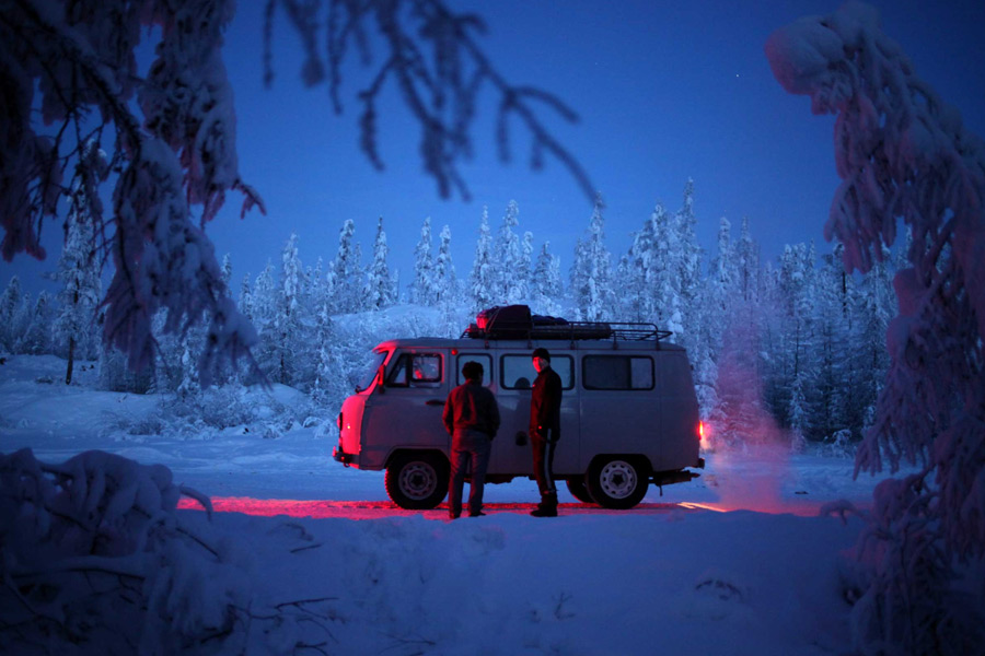 Photo of two people standing outside an old van at night time, in the snow. There is a blue filter over the whole image and it looks very cold.